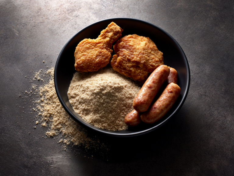 Flat lay on dark steel showing soy powder, pieces of golden fried chicken, and cooked plant-based sausages, representing diverse food applications